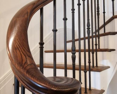 Walnut-toned curved handrail with scroll end over black metal spindles on a curved staircase